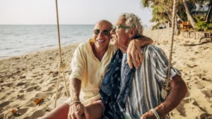 Happy senior gay man sitting with arm around boyfriend on rope swing at beach