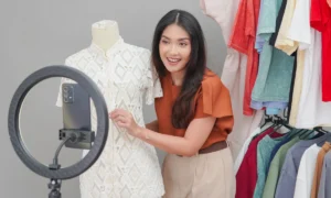 Young Asian woman doing live stream online shopping presenting a white dress with various clothes collection behind her with a happy expression.