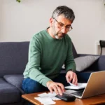 Middle aged man calculating expenses and managing the budget while using a calculator and laptop at home