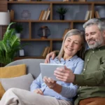 Happy senior couple relaxing on a comfortable sofa in their modern living room, smiling while browsing content on a digital tablet together, connectin