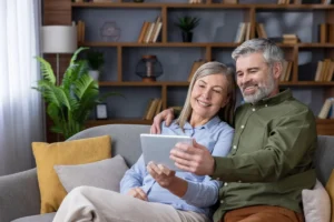 Happy senior couple relaxing on a comfortable sofa in their modern living room, smiling while browsing content on a digital tablet together, connectin