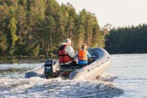 Elderly man and young boy in life jackets navigating an inflatable motorboat across a lake, with a forested shoreline in the background on a clear summer evening