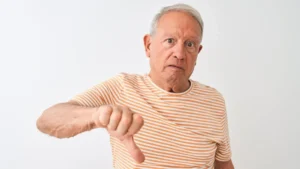 Senior grey-haired man wearing striped t-shirt standing over isolated white background looking unhappy and angry showing rejection and negative with thumbs down gesture. Bad expression.